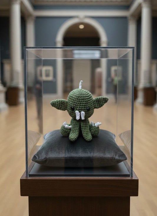 Crocheted Chupacabra figure on a pillow in a glass display case with a blurred museum setting.